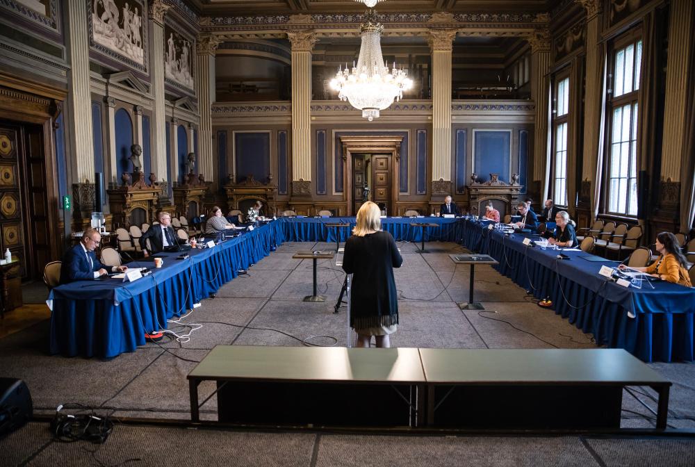 Säätytalon sali 15 jossa suuressa puolikaarteessa isoin turvavälein istuvat kokouksen panelistit. Paneelin moderaattori seisoo salin keskellä ja on selin kameraan.